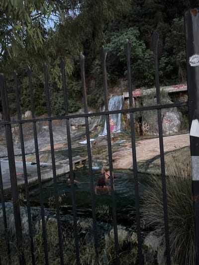 The image shows a fenced-off view of the Thermopyles Hot Springs in the Municipal Unit of Lamia, Greece. The viewpoint is through a metal fence, with greenery and trees visible on the left and behind the fence. The springs consist of multiple pools, with people swimming and relaxing in them. Water flows over a series of rocks, creating small waterfalls. Graffiti is visible on some of the rocks.