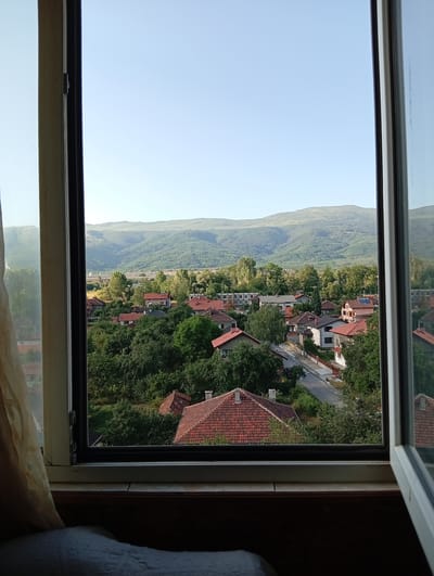 This is a photograph taken from inside a building, looking out through an open window. The window frame, painted white, is visible on the left, right, and bottom of the image. A cream-colored curtain hangs on the left side of the window.

The scene outside depicts a village or small town nestled amongst rolling green hills and a distant mountain range. The architecture features numerous houses with red tiled roofs, surrounded by lush green trees and vegetation. A road winds through the village, with several cars visible. The sky is a clear, pale blue, suggesting it is daytime, likely morning or late afternoon due to the soft light. The overall impression is one of a peaceful rural or suburban setting in Pirdop, Bulgaria. There are no people or specific activities clearly depicted, focusing instead on the landscape and the residential area. The image evokes a sense of calm and tranquility, offering a glimpse into a serene, sunlit day in a quiet community.