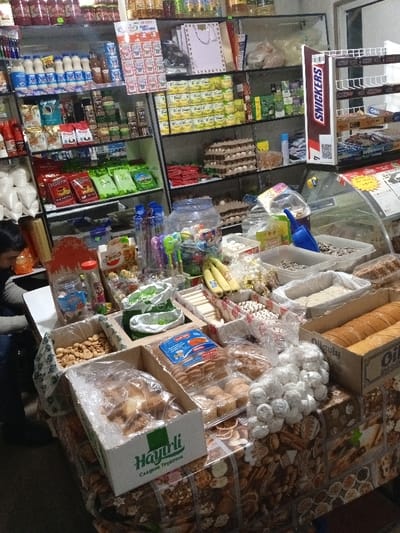 This image captures the bustling interior of a small grocery store or market stall in Avval', Uzbekistan. The scene is filled with a wide assortment of products, displayed both on shelves and across a prominent counter.
On the left, a person with dark hair, wearing a dark top, is partially visible from the side, possibly browsing items or attending to the store.
The background features shelves stocked with various packaged goods. These include rows of bottled beverages like milk and juices, cooking oils, bags of coffee or tea, jars, canned foods, and spices. Several stacks of eggs in cardboard cartons are also visible on the shelves. A display stand for "SNICKERS" chocolate bars is prominent on the right side of the shelves, alongside packaged "Oreo" cookies and other snacks.
The foreground is dominated by a counter laden with an extensive selection of bulk items. These are presented in open bins, plastic containers, and bags. Visible items include various nuts, dried fruits, fresh bananas, sunflower seeds (one bin contains a blue scoop), and an assortment of baked goods such as cookies and pastries. A bag of white, sugar-dusted candies is also present. Several cardboard boxes are used for displaying products; one box clearly shows the brand "Hayirli" and "Сладкие трубочки" (Sweet tubes/rolls) in Cyrillic script. Another box is partially visible with "Oil" printed on it. The counter is covered by a patterned tablecloth featuring images of nuts and various baked treats.
The setting suggests a well-stocked local shop catering to daily needs, reflecting a typical retail environment in Central Asia. The mixture of international brands with local or regional products, indicated by the Cyrillic text, aligns with the specified location context. The lighting suggests an indoor environment, with no discernible cues for the time of day or outdoor weather. No specific activity beyond shopping or retail is depicted, and no strong emotions are visible.
