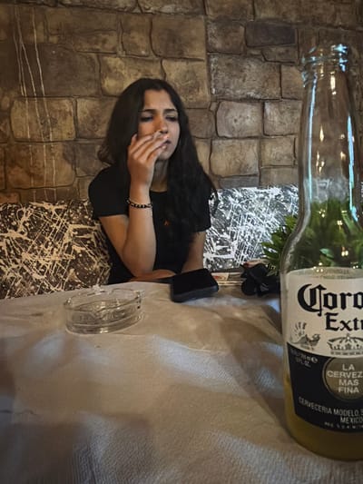 A young woman with dark, wavy hair is seated at a table in what appears to be a restaurant. She is wearing a black t-shirt and is holding a cigarette to her lips, with smoke emanating from it. On the table in front of her is a glass ashtray. To the right of the frame, a bottle of Corona Extra beer is visible, with a plant behind it. The background features a stone wall. The overall setting suggests an indoor dining or drinking establishment, likely in the city of Kermen, Bulgaria.