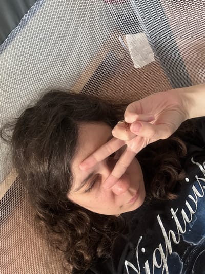 A person is lying down, with their face tilted upwards and to the right. They have dark, curly hair that falls around their shoulders. Their eyes are closed, and they are holding up their right hand to their face, forming a peace sign with two fingers extended over their forehead and cheek. A ring is visible on their ring finger. The person is wearing a dark t-shirt with white, stylized lettering that appears to read "Nightwish."
The person is positioned on a surface covered by a fine white mesh netting, which also partially obscures a wooden surface underneath. A small white tag is visible attached to the mesh in the upper right quadrant of the image. The overall environment appears to be an indoor setting, possibly a room or a portable crib, with warm lighting. The image is a close-up, taken from a high angle looking down at the person. The specific location in the city of Červená Píska, Czechia cannot be determined from the visual information provided.