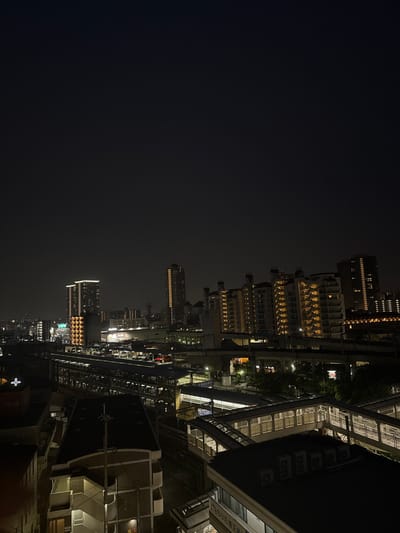 The image is an aerial view of the city of Takarazuka, Japan, taken at night from a rooftop. The coordinates provided, 34.811 latitude and 135.342 longitude, correspond to a location within Takarazuka. The scene is dominated by a cluster of tall, lit apartment buildings in the center-right of the image. These buildings are arranged in a seemingly random pattern and are partially obscured by dark, leafy trees. In the foreground, there are several smaller, low-rise buildings, some of which have illuminated windows. A network of elevated walkways and platforms stretches across the image, seemingly part of a pedestrian bridge system. Several rows of lights, most likely street lamps, can be seen running along the edges of the walkways. In the distance, a multitude of other lights are visible, suggesting a sprawling urban environment. The sky is dark and almost completely obscured by a thick layer of clouds or haze. The scene is devoid of any people or vehicles.  There is no sign of any activity in the image, indicating a late-night or early-morning time.