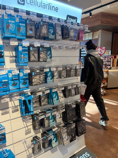 This image captures an indoor retail environment, likely in Dorno, Italy, showcasing a person browsing electronic accessories. The scene is brightly lit, with natural light creating distinct shadows on the light-colored tiled floor, indicating it is daytime.
In the foreground, a display rack filled with numerous packaged electronic accessories from the brand "cellularline" is prominent. The products include various types of headphones, earbuds, and phone mounts such as "SLIDE", "DASH PAD PRO", "HUG", and "SPIN MIRROR". Other product lines like "COLOR", "EAGLE", and "FIT" are also visible. These items are presented on white metal grid shelves with clear plastic hooks, each bearing small white price tags.
A person, seen from behind and slightly to the side, is leaning forward, attentively examining the products on the left side of the display. They have dark, curly hair, possibly tied back, and are dressed in a dark leather jacket over a grey hooded sweatshirt, dark pants, and light-colored sneakers.
In the background, beyond the person and the accessory display, other merchandise shelves are visible. A red tag with "ty" on it, commonly associated with Ty Beanie Babies, and several small plush toys can be seen, suggesting a diverse retail establishment, possibly a gift shop or general store. A large, illuminated sign on the left clearly displays "cellularline" and "travel".
Visible text includes: "cellularline", "travel", "SLIDE", "COLOR", "DASH PAD PRO", "SPIN MIRROR", "HUG", "EAGLE", "FIT", "ty", "FOR ALL SMARTPHONES", "C", "V", "Va", "owe", "CC", "Θ", "0", and the partial "SUC". The overall activity is a person casually shopping or browsing.