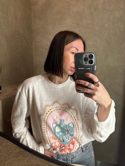 An adult female is captured in a mirror selfie within an indoor setting in Усады, Russia. She has dark, shoulder-length bob-style hair with subtle lighter highlights, and fair-to-medium skin tone with visible minor facial irregularities on the left cheek/forehead. Her lips are slightly parted, showing a natural or lightly glossed finish. She wears a loose-fitting, long-sleeved white t-shirt featuring a prominent circular graphic design. The graphic includes an urn-like vessel in shades of blue and teal, a starburst motif, and patterns in pink, orange, and red tones; reversed text, possibly "Cielo Olojo" or "Cielo d'Olio," is visible above the design. Light-wash denim jeans are partially visible below the shirt.
The subject holds a dark-cased smartphone (appears to be an iPhone with a triple-lens camera array) in her left hand, positioning it to capture her reflection. The phone case has reversed text, possibly "VVAA" or "DVVA." On her left hand, she wears two rings: a wide, multi-banded silver ring on the ring finger, and a single silver band with a small, dangling flower charm on the middle finger.
The background consists of a textured wall, finished in a light brown or greige color with subtle, swirling plaster patterns. A corner of the room is visible. To the upper left, a small section of a light-colored wooden shelf or counter is present, holding a dark, slender bottle, possibly a diffuser. The bottom of the image shows a dark, horizontal line, indicative of the edge of the mirror frame or a counter surface reflecting the subject's lower body. The lighting is diffused and even, with no harsh shadows.