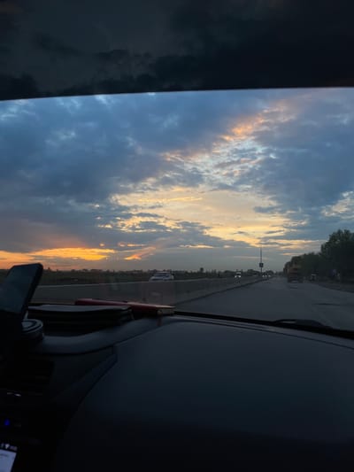 The image shows a view from inside a vehicle, likely a car, driving on a road in Sanchikul, Uzbekistan. The focus is on the sunset visible through the windshield, with partly cloudy skies displaying hues of blue, yellow, and orange. The road stretches forward, with other vehicles and infrastructure like street signs and vegetation visible in the distance. The dashboard and parts of the interior of the vehicle are visible in the foreground.