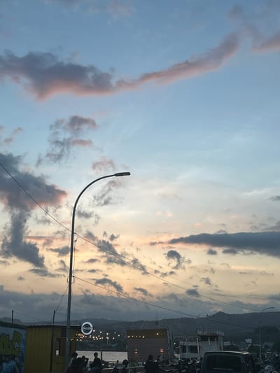 The image depicts a street scene at dusk in Bantaeng, Indonesia. The sky is filled with a mix of gray and pinkish clouds, illuminated by the setting sun, with a pale blue sky above. A tall, curved street lamp is prominent in the foreground, with electrical wires crisscrossing the frame. In the mid-ground, there are several low-rise buildings, one yellow structure with graffiti on its side, and another with a circular sign displaying the word "Hagemony." String lights are visible across the buildings. A body of water, likely a bay or harbor, can be seen behind the buildings. The foreground is populated by a group of people, some seated, and several motorcycles. In the background, hills and mountains form the horizon. The overall atmosphere suggests a lively evening gathering or marketplace by the waterfront.