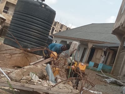 The image depicts a scene in Achara Ndiagu Omegio, Nigeria. A man wearing a blue shirt is working amidst construction materials. A large, black plastic water tank is prominently visible. There are several yellow containers, a metal bucket, and exposed rebar scattered around. The background shows unfinished buildings and houses. A tin roof covers one of the structures. The environment appears dusty.