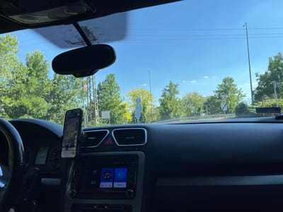 This media file is a first-person perspective shot from inside a car, looking out at the road and dashboard. The scene is set on a sunny day, with a clear blue sky and lush green trees lining the road. The car's dashboard is visible, including the steering wheel, instrument cluster, air vents, and a central infotainment screen displaying music and phone icons. A smartphone is mounted on the dashboard, displaying a navigation app with a map and route information. The time appears to be daytime, likely late morning or early afternoon, given the bright sunlight. The location cues, such as the road, trees, and a lamppost, suggest an outdoor setting, possibly a suburban or rural area. There are no people visible in the frame. The overall impression is one of a leisurely drive on a pleasant day. The visible text on the infotainment screen includes "21 km/h" and what appears to be a Bluetooth connection icon. The navigation app on the phone also displays text, but it is not clearly legible.
