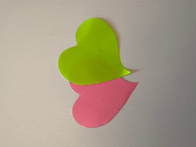 The image shows two heart-shaped cutouts, one lime green and the other pink, placed on a textured, light-colored surface. The lime green heart is positioned above and slightly to the left of the pink heart, with their tips overlapping. The background surface appears to be a light gray or beige, with a subtle, speckled texture and faint horizontal lines. There are no people or animals visible, and no clear indication of a specific time of day, weather, or location cues beyond the general indoor setting. The image focuses solely on the two heart shapes, creating a simple and graphic composition. There is no visible text in the image.
