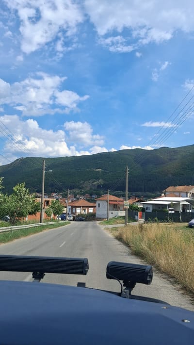 This is a vertical, eye-level, outdoor, daytime photograph taken from the perspective of someone inside a vehicle, looking forward and slightly upwards. The foreground shows the hood of a dark blue vehicle with two large, rectangular LED light bars mounted on it. The road ahead is paved and appears to be a country road or a road within a small town. To the left of the road, there is a metal guardrail and green foliage. On the right side of the road, there is dry, tall grass.
In the mid-ground, the road leads into a village. Several houses with red-tiled roofs are visible. Some houses are partially constructed, showing exposed brickwork. There are utility poles with overhead wires lining the road.
In the background, a large, densely forested green mountain dominates the scene. The sky above is a clear blue with scattered white cumulus clouds. The overall impression is of a rural or semi-rural setting in a mountainous region. The location is identified as Mosomishte, Bulgaria. There are no people or animals clearly visible in the image. The scene depicts a quiet, possibly residential area on a sunny day.