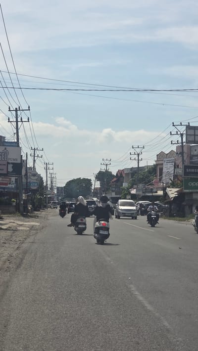 The image depicts a street scene in Barat Yaman, Indonesia, with a road leading into the distance. Several scooters are riding on the road. Various buildings and signage are visible along the roadside, including advertisements for Samsung, Oppo, Xiaomi, and Hannochs. Overhead power lines run along the street. The sky is blue with scattered clouds.