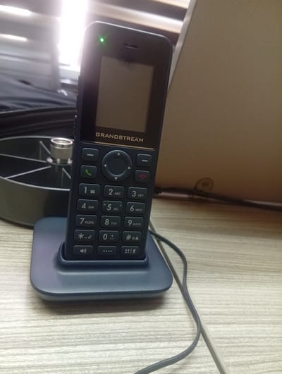 A close-up, eye-level, indoor shot shows a black cordless Grandstream telephone resting in its dark blue charging base on a light-colored wooden desk. The telephone has a small, dark screen at the top, a green indicator light illuminated above the screen, and a standard keypad below. The keypad features numbered buttons with corresponding letters, a navigation dial, and soft keys. A power cord is plugged into the back of the charging base and runs out of frame to the right. In the background, to the left, a black object with a ribbed edge is visible, and behind it, bright light suggests a window. To the right, a tan, textured surface, possibly a wall or partition, occupies the background. The lighting is bright, with a strong glare coming from the left, indicating overhead lighting or natural light from a window. The scene is static and appears to be a typical office or home environment. The image was taken in Shiroro, Nigeria.