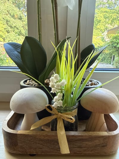 This image shows a wooden tray on a windowsill, containing two decorative mushroom figures, a glass jar with faux lily of the valley, and two potted plants with dark green leaves. The tray is placed on a light-colored wooden surface. Through the window, a blurry background of green trees and foliage is visible, suggesting an outdoor setting. The scene is lit by natural light, casting soft shadows. No people or animals are present. The location is not specified in the image, but the prompt implies it is in Herford, Germany.