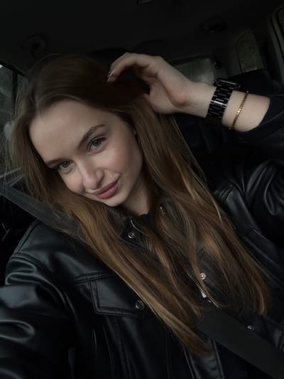 A female individual is depicted seated in the passenger side of a vehicle in Stupovi, Montenegro. She has long, light brown hair parted on the left, and light-colored eyes. Her head is tilted slightly, and she displays a subtle smile, looking directly at the viewer. Her left hand, with light-tipped fingernails, is raised and touches her hair above her right ear. On her left wrist, she wears a black metal link watch and a thin gold-colored bracelet composed of small, flat circular elements. She is dressed in a black leather-style jacket over a dark inner garment. A black seatbelt is visible, extending diagonally from her right shoulder across her chest and torso. The vehicle's interior serves as a dark, indistinct background, with faint indications of an exterior view through the windows.