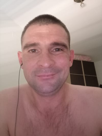 The media file is a close-up, head-and-shoulders selfie of a man with short, dark hair. He is wearing earbuds, with one earbud visible in his left ear and its wire extending downwards. The man is shirtless, revealing his shoulders and upper chest, which has some light hair. He has brown eyes and appears to be smiling faintly. The background is a lightly colored wall with what looks like a ceiling light fixture at the top. To the right of the man, a section of a dark brown wardrobe with four square doors is visible, topped by a pink and white patterned object, possibly a pillow or blanket. The lighting suggests it is daytime indoors. There is no visible text in the image. The man's expression suggests a relaxed or content emotion.