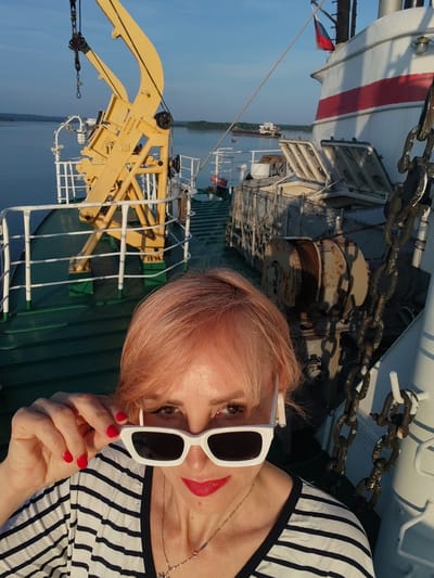 A medium shot captures a woman with pink hair, wearing white cat-eye sunglasses and a black and white striped shirt, on the deck of a boat. She is holding her sunglasses with her right hand, revealing her eyes and red lipstick. Her left hand is not visible. The background shows the deck of a boat with a yellow crane and various metal structures, including chains and winches. The boat is on a body of water, with land and other boats visible in the distance under a clear sky. The setting is the city of Shchetinino, Russia.