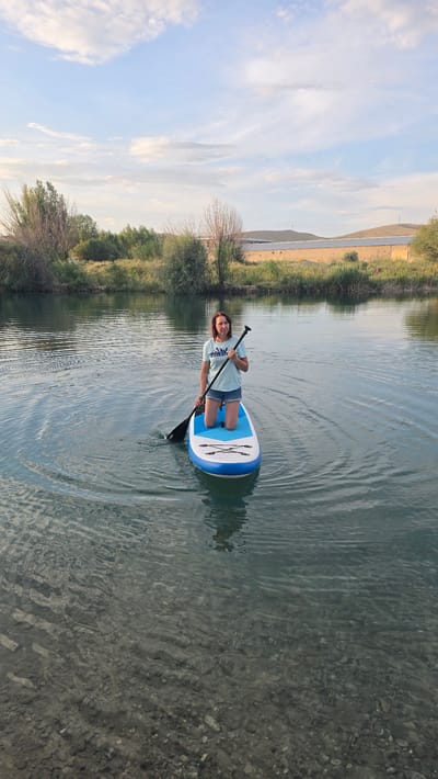 A woman is kneeling on a blue and white paddleboard in a body of water. She is holding a black paddle with a black blade. The paddleboard is in the middle of the water, with ripples spreading out from it. The woman is wearing a light blue t-shirt with a graphic on it and denim shorts. She has brown hair and is looking at the camera. The water is clear and shows pebbles on the bottom. In the background, there is a shoreline with green vegetation and trees. Beyond the vegetation, there are hills and buildings, possibly solar panels. The sky is partly cloudy with a mix of blue and white. The scene appears to be outdoors in a natural setting.