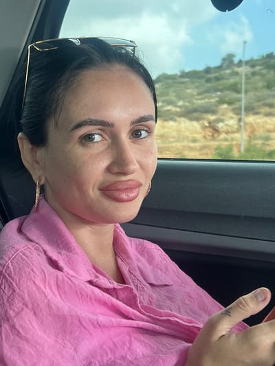 The image shows a close-up of a woman in a car, facing the camera. She has dark hair pulled back and is wearing large, hoop earrings and a pink collared shirt. Sunglasses are perched on top of her head. Her lips appear plump. The background visible through the car window suggests a rural or semi-rural setting with hills and vegetation, consistent with locations in the Galil Tachton Regional Council, Israel. The lighting indicates it is daytime. A portion of the car's interior, including the door panel, is visible. The woman's right hand is partially visible in the foreground, showing a tattoo on her thumb.