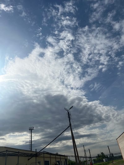 The image shows a daytime view in Бизяки, Russia, looking upwards at the sky. A mix of cirrus and cumulus clouds is visible, creating a varied cloudscape. The sky transitions from a light blue to a darker, more overcast gray. Utility poles and wires extend into the frame from the right side, with a building partially visible in the lower right corner. A transmission tower is also seen among the electrical infrastructure.