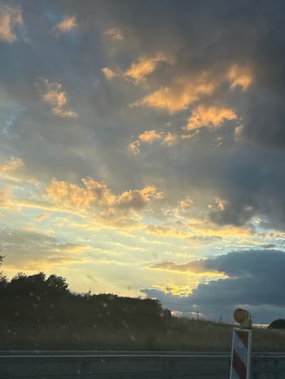 The image depicts a dramatic sky with scattered clouds illuminated by the golden hues of sunset or sunrise. The clouds on the left are darker, possibly indicating a storm, while those on the right are brightly lit, creating a stark contrast. The setting appears to be along a road or railway line, with visible tracks and a warning sign with red and white stripes in the upper right portion of the frame. A light fixture, possibly a signal lamp, is also visible near the sign. Silhouetted trees line the horizon, suggesting a rural or semi-rural location. The overall mood is one of natural beauty and possibly the calm before or after a storm. The time of day is clearly indicated by the warm, low-angle light casting long shadows and illuminating the clouds. There is no visible text in the image.