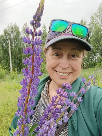 A woman is captured in a selfie in a field of lupine flowers. She is wearing a green hooded jacket, a patterned cap, and sunglasses with colorful reflective lenses. She is holding a bouquet of purple lupine flowers. The background shows a field of grass and trees. The location is identified as Gribchikha, Russia.