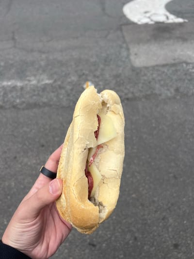 A hand holds a sandwich in an outdoor setting. The sandwich, made with a pale, crusty roll, contains cheese and sliced sausage. The hand, likely male, wears a black ring with gold accents. The background appears to be a paved surface, possibly a sidewalk or street, with a white painted marking visible in the upper left corner. The location is La Aldea de San Nicolás, Spain.