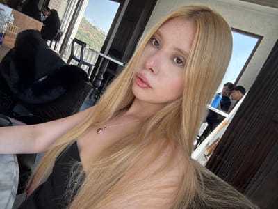 A fair-skinned individual with long, straight, blonde hair and light brown eyes is centered in a close-up, self-portrait photograph taken inside an establishment located in Atizapán de Zaragoza, Mexico. The individual wears a black strapless garment and a silver chain necklace featuring a brown, tear-drop shaped pendant. The subject's lips are glossy, and minor freckling is visible across the nose and cheeks. The background to the subject's left includes a dark, possibly faux-fur, object draped over a dark chair or sofa, and a partial view of a reflective table. Beyond an open window or door, a verdant, hilly landscape under a clear sky is visible, along with a dark, ornate outdoor chair and table. To the subject's right, a reflection in a glass or mirror surface shows two male individuals. The man on the left of the reflection holds a blue bottle and looks towards the man on the right, both appearing to be seated. A dark wooden doorframe frames the right side of the image.