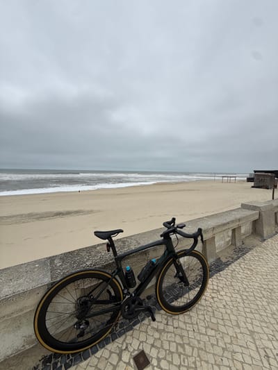 A dark-colored road bicycle with drop handlebars and two water bottles (one light blue, one black) leans against a weathered concrete wall. The bicycle features deep-section wheels with tan-colored sidewall tires, a red rear light, and a cycling computer mounted on the handlebars. The concrete wall separates a cobblestone promenade, composed of irregular light and dark grey stones, from a sandy beach. The wide, light-colored sand shows numerous footprints and extends towards the ocean. In the distance, a small, indistinct human figure is visible on the beach near the water's edge. To the right on the sand, a small wooden hut or shack and a separate wooden framework are present. The Atlantic Ocean, characterized by choppy, grey-green water and white breaking waves, stretches to the horizon. The sky is uniformly grey and heavily overcast, indicating a cloudy day at Praia de Mira, Portugal.