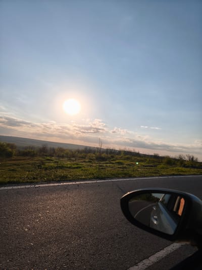 The image shows a view from inside a moving car. The location is identified as Zdravets, Bulgaria, based on contextual clues or external information not visible in the image itself.

The image captures the following elements:

*   **Environment:** The external environment consists of a field with low vegetation and some sparsely distributed trees. The sky is visible, with a bright sun and some scattered clouds.
*   **Infrastructure:** A section of an asphalt road is visible in the foreground. A white road marking line separates the shoulder from the lane.
*   **Vehicle:** A car's side mirror is prominently featured in the lower right corner, showing a reflection of the road behind and part of the car's body. The car is presumably in motion.
*   **Light:** The scene is brightly lit by the sun, casting shadows and creating highlights on the road surface.