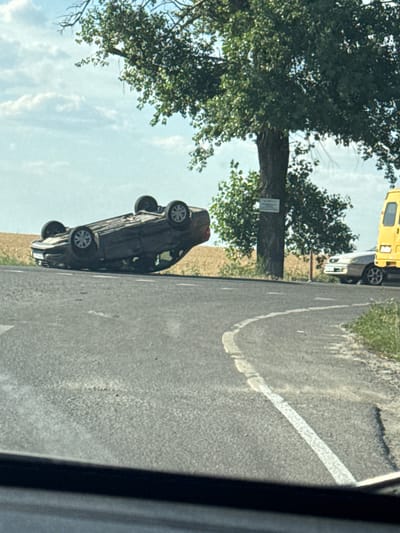 In Varenovka, Russia, a car accident has occurred. A dark-colored car is overturned on its roof on the side of a paved road. The car is positioned on the right side of the road, with its wheels in the air. The road appears to be in a rural or semi-rural setting, with a field of dry vegetation visible in the background. A large tree with green foliage stands near the road. There is also a yellow vehicle, possibly a van or truck, parked on the right side of the road, behind the overturned car. Another car, a light-colored sedan, is parked to the left of the yellow vehicle. The sky is partly cloudy. The image is taken from a low angle, possibly from another vehicle.