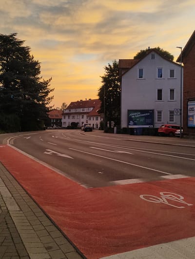 The image depicts a street scene at dusk in Göttingen, Germany. The sky is filled with soft, warm hues of orange and yellow, indicating the setting sun. In the foreground, a paved sidewalk runs alongside a road marked with white directional arrows. A designated bicycle lane, painted in bright red, is clearly visible, featuring a white bicycle symbol.

On the right side of the street, a white building with multiple windows stands prominently. A large billboard on this building reads "JÜDISCH IN GÖTTINGEN" (Jewish in Göttingen). Adjacent to the billboard are blue trash bins. Further down the street, residential buildings with tiled roofs can be seen, along with parked cars. A red car is parked in front of the white building, and a silver car is parked behind it. A speed limit sign (30) is visible on a pole near the cars.

To the left of the road, dense, dark green trees create a strong contrast with the illuminated sky. The overall impression is a quiet urban setting during twilight, with clear indications of infrastructure for both vehicular and bicycle traffic.