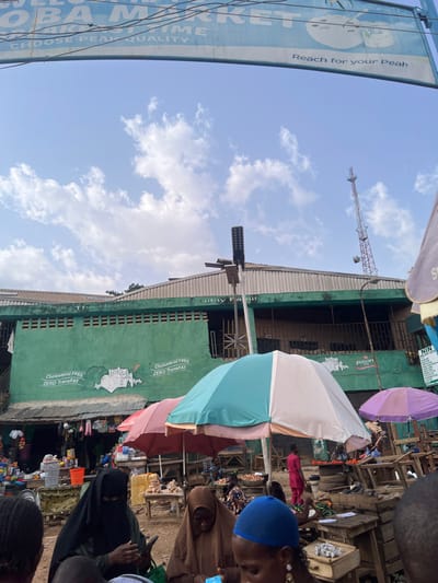 The scene takes place in an open-air market located at the coordinates 7.253, 5.195 in the city of Ondo, Nigeria. There are multiple stalls under colorful umbrellas. A group of people are engaged in buying and selling produce. Two women in hijabs are interacting, one is holding a phone. There are multiple objects in the market, including wooden tables, chairs, and baskets filled with produce. The sky is bright blue with scattered white clouds. A tall cellular tower is visible in the background. A sign above the market reads "Oba Market. Choose Peak Quality. Reach for your Peak." There is another sign on the wall of the market reading "Cholesterol FREE ZERO TransFAT."