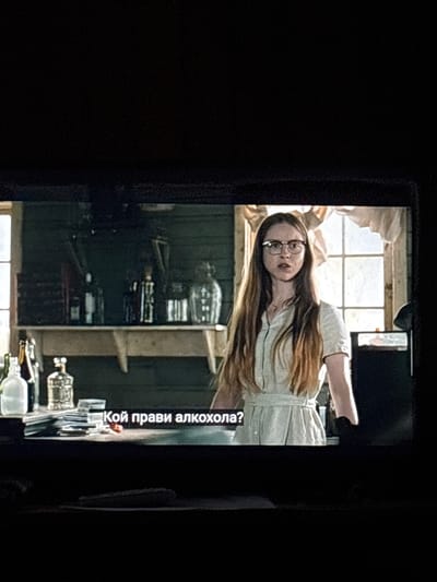 An image, originally captured in portrait orientation and rotated 90 degrees, depicts a young woman indoors. She has long brown hair, wears glasses, and is dressed in a light-colored, possibly off-white or light grey, short-sleeved top or dress. Her expression is serious and direct as she looks towards the viewer, with her right arm slightly raised and extended in a gesturing manner.

Behind the woman, a large window with multiple panes is visible, through which bright daylight streams, indicating it is daytime. Below the window, a counter or shelf holds several clear and dark glass bottles, including a prominent large carboy and smaller bottles with stoppers, suggesting an area for brewing, distilling, or storing alcoholic beverages. The room's interior features dark-colored wooden paneling on the walls, contributing to a rustic or workshop-like atmosphere.

Overlaid on the bottom right of the image (when correctly oriented) is Bulgarian text that reads: "Кой прави алкохола?". This translates to "Who makes the alcohol?". The scene captures a moment of inquiry or observation related to the activity suggested by the bottles, set in what appears to be a rural Bulgarian location like Brenitsa.