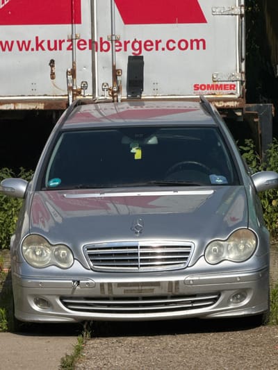 A silver Mercedes-Benz C-Class station wagon is parked on a paved surface, likely a street or parking lot, in Schkopau, Germany. The car is positioned directly in front of a large white trailer with red lettering on its side. The trailer displays the website address "www.kurzenberger.com" and a logo for "SOMMER ROAD CARGO SOLUTIONS". The car has its headlights on, and the front bumper shows some scuff marks and a tow hook attached. Green foliage is visible behind and to the sides of the vehicle. The car has a blue sticker on the windshield and a yellow air freshener hanging from the rearview mirror.