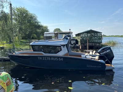 A motorboat is moored in calm, dark water near a wooden dock. The boat is dark blue with a light grey cabin and wooden accents. It has a Suzuki outboard motor at the stern. In the background, there are trees, a building with a camouflage covering, and other structures on the shore. The sky is clear blue. The boat has the registration number TH 2178 RUS 64 and the model name VOYAGER 600. The location is identified as General'skoye, Russia.