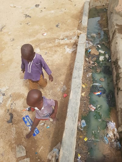 Two young boys are sitting on a sandy, littered ground next to a concrete drainage ditch filled with stagnant water and trash. The boy on the left is wearing a purple long-sleeved shirt and brown pants. The boy on the right is wearing a short-sleeved shirt and shorts. There is a toy gun near the boy on the right. The surrounding area is littered with debris, including plastic wrappers and paper. The photo appears to be taken from directly above, looking down on the scene.
The location is implied to be in Dutsinma A, Nigeria, based on the prompt.