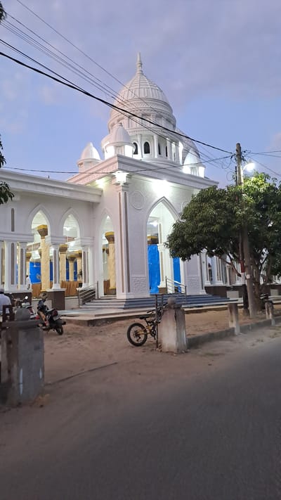 The image depicts a white building with multiple domes and arches, likely a mosque, located in the city of Lhokawe Teungoh, Indonesia. The building features intricate architectural details, including decorative carvings and columns. In front of the building, there is a paved area with several parked motorcycles and a bicycle. A large tree stands to the right of the building, and utility poles with overhead wires are visible in the background. The sky is partly cloudy, indicating it is either early morning or late evening. The overall scene suggests a public space within a community, possibly a religious or cultural center.