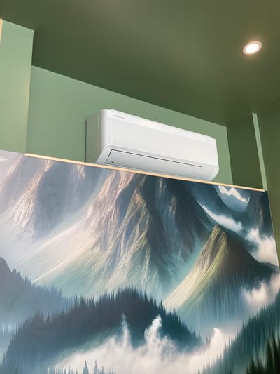 An indoor scene features a white Samsung air conditioner unit mounted high on a light green wall, above a large, horizontally oriented artwork depicting a misty mountain landscape with a dense evergreen forest in the foreground. The ceiling, painted a dark green, has a recessed circular light fixture. The image provides no information about Meaux, France.