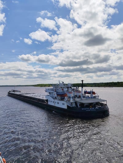 A barge, identified as "VOLGO-DON 5051" from Nizhny Novgorod, is seen on a body of water under a sky with scattered clouds. The barge is dark colored and appears to be a river transport. The background shows a shoreline with green trees. The water is dark and slightly rippled. The image does not appear to be located in Popovo, Russia.
