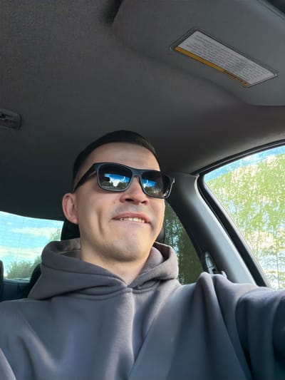 A person wearing sunglasses and a gray hoodie is in a car. The car's sun visor with a warning label is visible. The sunroof is open, revealing trees outside. The location is indeterminable.
