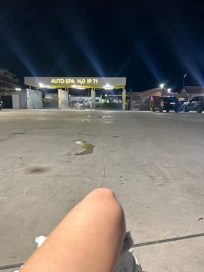 The image depicts an outdoor scene at night in what appears to be a car wash facility. A sign reading "AUTO SPA H2O IP 71" is visible, suggesting a possible business name and location near Plovdiv, Bulgaria. The foreground features a person's arm and possibly a part of their foot. The ground is concrete, marked with stains. Further back, cars are lined up at the car wash area, indicating activity at the facility. The sky is dark and some buildings are visible in the far background.