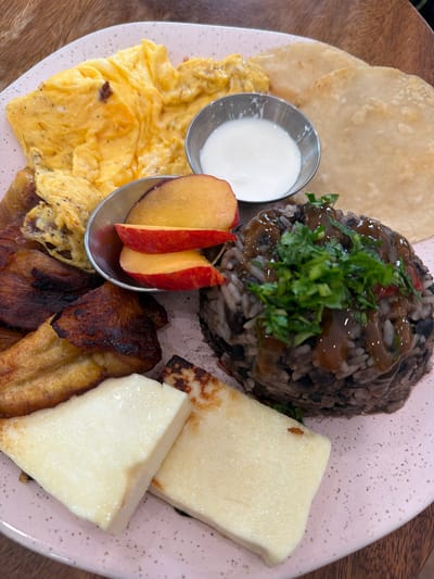 This is a close-up, top-down shot of a plate of food, likely a breakfast or brunch dish. The plate is pink with scattered dark speckles. The food includes several distinct components arranged on the plate. In the upper center is a mound of rice and beans, topped with chopped green herbs and a drizzle of dark sauce. To the right of the rice and beans are several rectangular slices of white cheese, some showing light browning from grilling. In the lower left quadrant, a pile of fluffy, yellow scrambled eggs is visible. Adjacent to the eggs, in a small silver bowl, is a white creamy substance, likely sour cream or yogurt. Next to the bowl of cream and partly overlapping the eggs is a small serving of sliced fruit, appearing to be peaches due to their red skin and yellow flesh. In the lower central and right portions of the plate are several pieces of fried plantains, some appearing caramelized and darker brown. To the left of the rice and beans and above the eggs are two thin, round, light-colored flatbreads, possibly tortillas. The overall presentation suggests a savory meal with elements of sweetness from the fruit and plantains. The lighting is bright, indicating daytime. The setting appears to be a restaurant or home dining table, with a wooden surface visible in the background. No people are visible. No text is discernible.