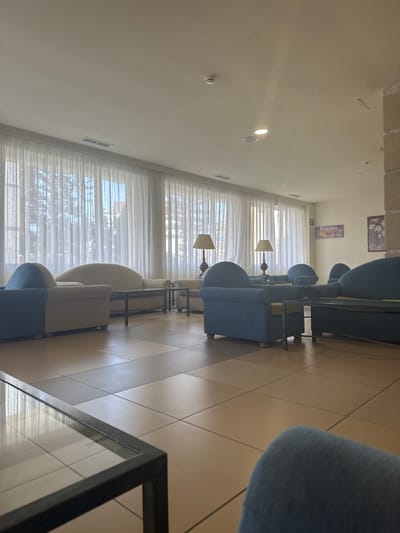 This image depicts the interior of a building, possibly a hotel lobby or a common area, with a view of white sheer curtains covering large windows. The room is furnished with several seating arrangements, including blue upholstered armchairs and sofas, and a cream-colored sofa. There are coffee tables in front of some of the seating areas, one with a glass top and metal frame, and another smaller one with a yellow cushion accent. Two lamps with beige shades are visible on side tables. The floor is tiled with a pattern of light brown and beige squares. On the wall in the background, a sign with text in Spanish, including "Planta" and numbers, is mounted. There are no people visible in the image. The lighting suggests daytime, with natural light filtering through the curtains. The overall impression is one of a clean and organized common space. The specific location within Fuengirola, Spain, cannot be determined from the visual information provided.