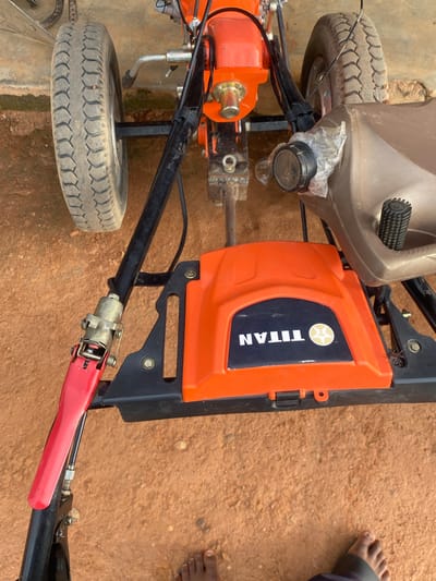 In Jere South, Nigeria, a close-up shot captures a portion of a tillering machine, possibly a walk-behind model, resting on reddish-brown soil. The machine features an orange engine cover with a black panel displaying the "TITAN" logo in white lettering and a star emblem. To the right, a brown plastic fuel tank is visible. The machine is equipped with black metal framing and a prominent black handle with a red lever mechanism. A thick black cable runs along the handle. One tire, with a weathered tread pattern, is visible on the left side of the frame. The background shows a textured surface, possibly a wall. A bare foot, belonging to a person not fully in frame, is visible at the bottom edge of the image.