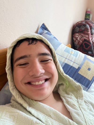 A close-up selfie of a young male with dark hair, wearing a light green hooded bathrobe, is taken from a slightly high angle. He is lying down, with a pillow patterned with blue and beige squares behind his head and to his right. His eyes are mostly closed, and he is smiling broadly, showing his teeth. His hair appears wet. Behind him, on the right side of the frame, a backpack with a red, black, and white abstract pattern is visible leaning against the wall, with two white cylindrical objects on top of it. The background wall is a textured, light pinkish-beige color. The wooden headboard of a bed is partially visible on the left. The scene is indoors, likely in a bedroom. There is no specific indication of the city of General Todorov, Bulgaria.