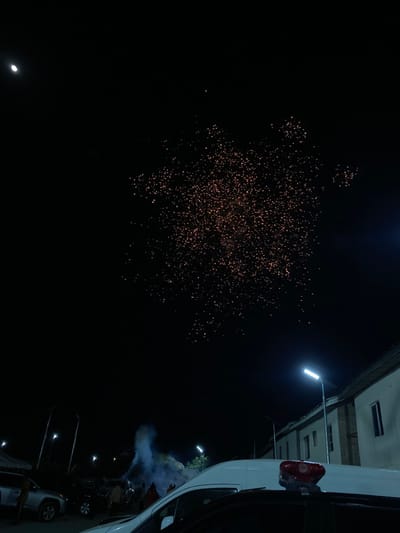 Fireworks explode in the night sky, illuminating the darkness with golden sparks. A street light shines near the upper right corner of the frame, illuminating a building and a white van. Below, on the ground, faint figures of people are visible, surrounded by smoke. Parked vehicles line the street, their lights adding to the overall illumination of the scene.