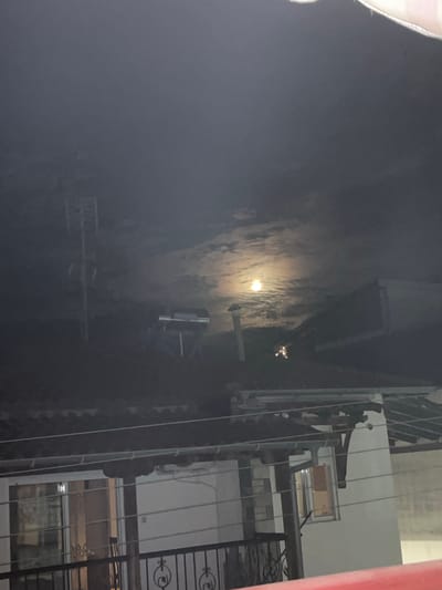 The image shows a night scene in Nea Flogita, Greece, with a bright full moon partially obscured by clouds. The moon is positioned in the upper center of the frame, casting a soft glow. In the foreground, several rooftops are visible, characterized by dark, tiled surfaces. A tall, metallic antenna or tower stands on the left side of the image. To the right of the antenna, a structure that appears to be a solar water heater or a similar apparatus is situated on a rooftop. A chimney or vent pipe is also visible on one of the roofs. Below the rooftops, the upper floors of residential buildings are partially seen, with illuminated windows suggesting indoor activity. A balcony railing with decorative metalwork is present in the lower part of the image, indicating a viewpoint from a higher elevation. The overall atmosphere is tranquil and slightly ethereal due to the moonlit sky and the dim lighting.