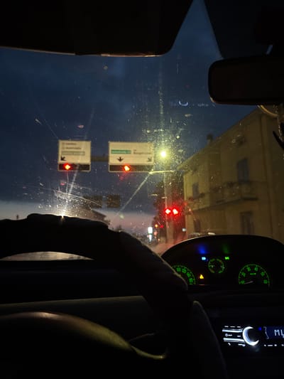 The image is taken from the interior of a car at dusk, looking forward through the windshield. The sky is a deep blue, transitioning to lighter hues near the horizon, with some clouds visible. A street light illuminates the scene from the right. Two green directional signs with white text and arrows are mounted on poles; one points left and the other right. Beneath these signs, red traffic lights are illuminated. A yellow building with windows and a balcony is visible on the right side of the frame. The dashboard of the car is in the foreground, showing a speedometer, tachometer, and other indicator lights. The steering wheel is partially visible, with a hand resting on it. The scene suggests the car is stopped at a traffic light. The digital display of the car stereo is visible, showing "ML" and other characters. The location is Mezzana Sant'Antonio, Italy.
