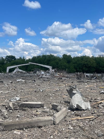 The image shows a field of debris in Terny, Ukraine (latitude: 50.979, longitude: 33.946). The debris is likely the result of a bomb blast. The debris is scattered across the field, with some large pieces of concrete, as well as smaller pieces of wood and metal. The sky is blue with white clouds. There are trees in the background. There are no people visible in the image.