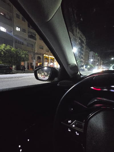 Inside a car at night, the steering wheel dominates the right side of the frame with its illuminated controls. Through the windshield, a dimly lit city landscape appears, featuring buildings and streetlights. The car's side mirror shows a more detailed view of a building with illuminated windows and architectural details. Trees and street vegetation are also visible. The overall scene conveys an urban nighttime drive.